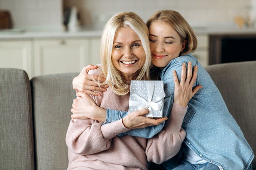 Surprise for mother's day or birthday. Loving young adult daughter giving a present to her beloved middle-aged caucasian mom sitting on the sofa in the living room | gifts for moms who doesn’t want anything