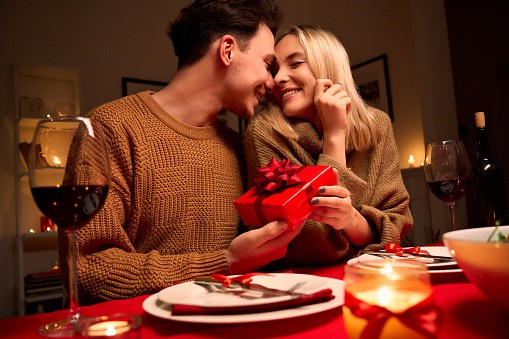 Happy young couple celebrating anniversary or Valentines day having romantic dinner at home table. Loving man giving red gift box hugging beloved woman making present surprise on date in candle light. | 14 Personalized Valentine’s Gifts for Him