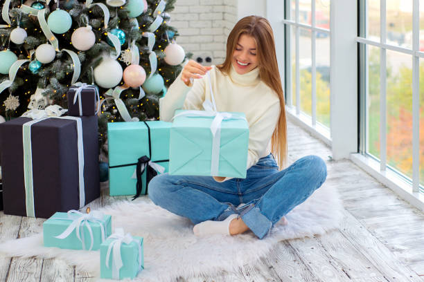 beautiful young 25 years old woman sits near a christmas tree and opens presents at christmas - gifts for a 25-year-old woman