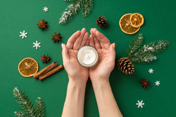 First person top view photo of woman's hands holding small jar with cream on palms pine cones dried orange slices cinnamon sticks anise snowflakes fir branches in frost on isolated green background - gifts for someone with sensitive skin