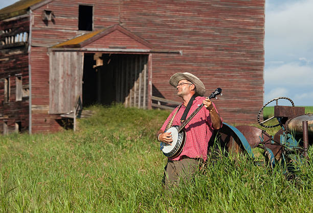 A man playing a banjo in a rustic rural scene. Abandoned grain elevator in distance. Horizontal colour image. White male. Early 40s. Prairie setting. - Gifts for banjo players