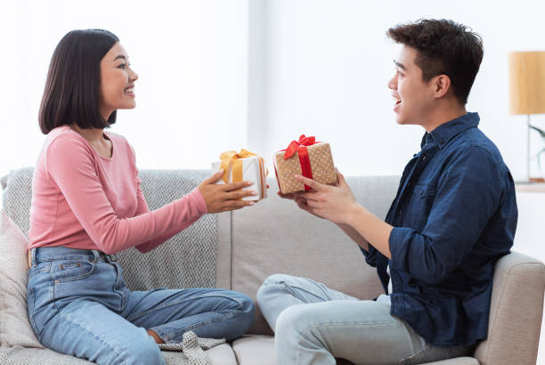 Valentine's Day Celebration. Lovely Asian Couple Giving Gifts Boxes Congratulating Each Other Celebrating Holiday Together Sitting On Sofa At Home. Romance, Holidays Presents Concept - gifts for Korean girlfriend