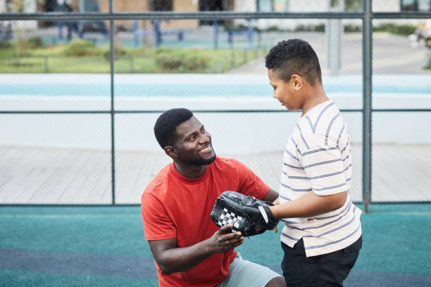 Happy handsome African-American father who acts as a baseball coach with beard chatting with teenage son in baseball glove while preparing him for game - baseball coach gifts