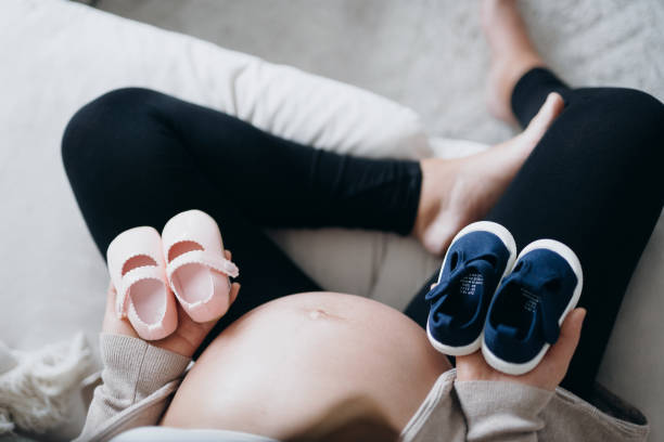 Overhead view of Asian pregnant woman holding a pair of blue and pink baby shoes in front of her belly. Expecting a new life, mother-to-be, gender reveal concept - gender reveal gifts