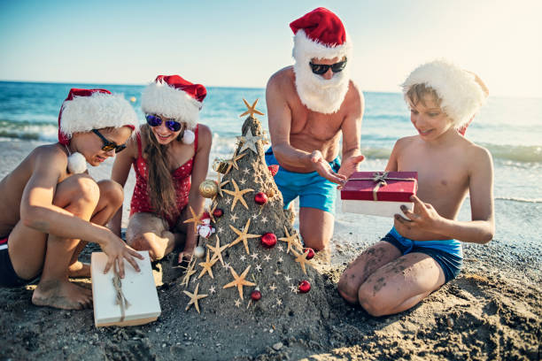 Father and kids are having fun during summer Christmas. Kids wearing Santa hats are opening gifts under sand Christmas tree. - gifts for beach lovers