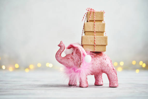 Holiday background with pink elephant wearing a Santa hat and carrying a stack of gifts for Christmas - elephant Mother's Day gifts