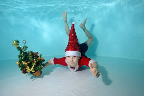 Cute little boy in Santa Claus costume underwater at the bottom of the pool. He holds a Christmas tree in his hands and gives a thumbs-up. Active child. Healthy lifestyle. Swimming lessons underwater - gifts for swimmers