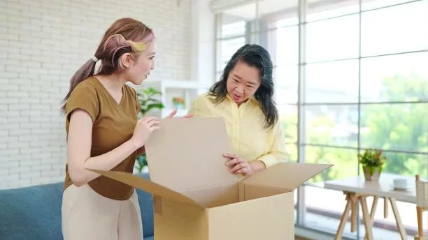 Happy young Asian daughter and mother looking into cardboard box, unpacking with feeling excited getting parcel with wished purchase item from internet store. Online shopping concept - gift for mom returning to work