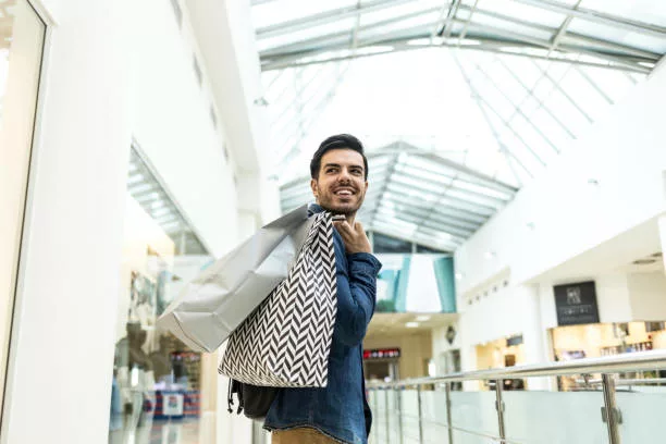 Beautiful young man holding paper bags and walking in mall, shopping. - gifts for the metrosexual man